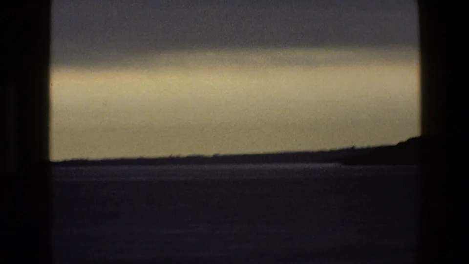 A low, flat horizon and pale hazy sky seen from behind a window in archival 8mm England footage.