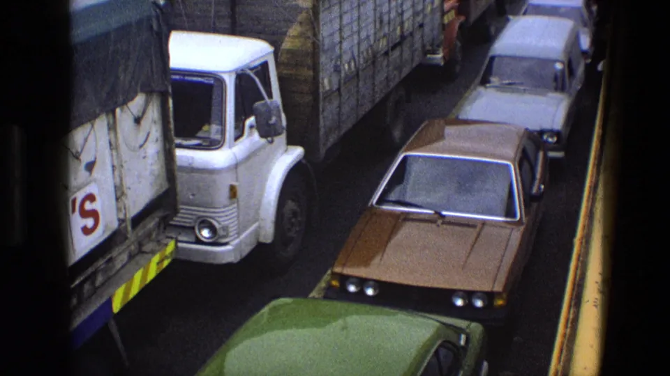 1969 England traffic jam with a white delivery lorry, brown sedan, green car, and tightly packed road vehicles.