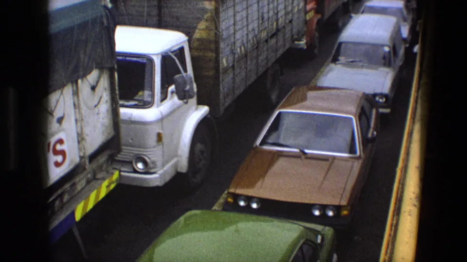 High-angle 1969 England traffic view with a white lorry, brown sedan, grey car, and green car roof packed in one lane.