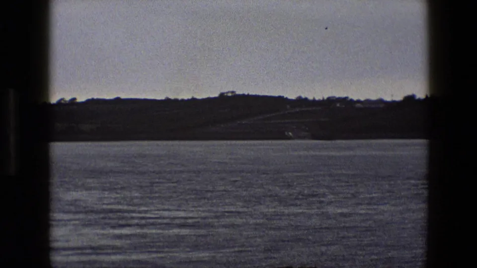8mm travel footage shifts from ferry vehicles to calm water and a low English shoreline in the distance.