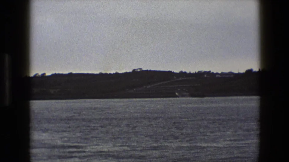1969 England 8mm frame showing open water, a far shoreline, low hills, and small distant buildings under a pale sky.