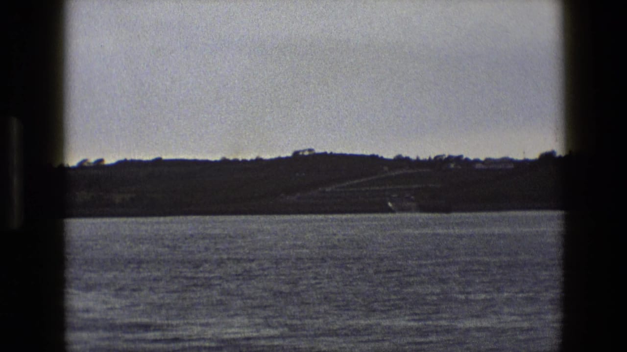 Muted 8mm view across water toward an English hillside, with small buildings and a road visible in the distance.
