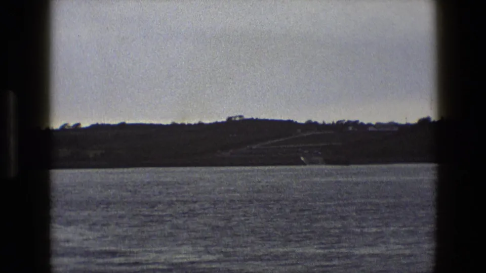 Muted 8mm view across water toward an English hillside, with small buildings and a road visible in the distance.