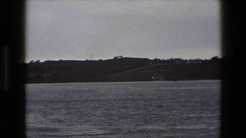 1969 England home-movie frame with rippled water, a distant hillside, and a small ramp or road descending near the shore.