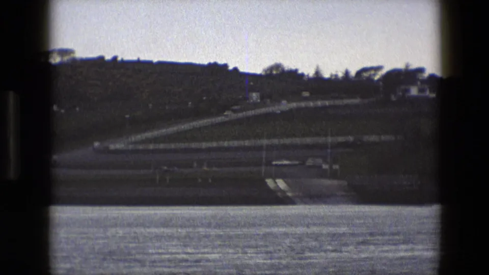 Zoomed 1969 England 8mm frame showing water, a shoreline ramp or road, hillside houses, and a few distant vehicles.