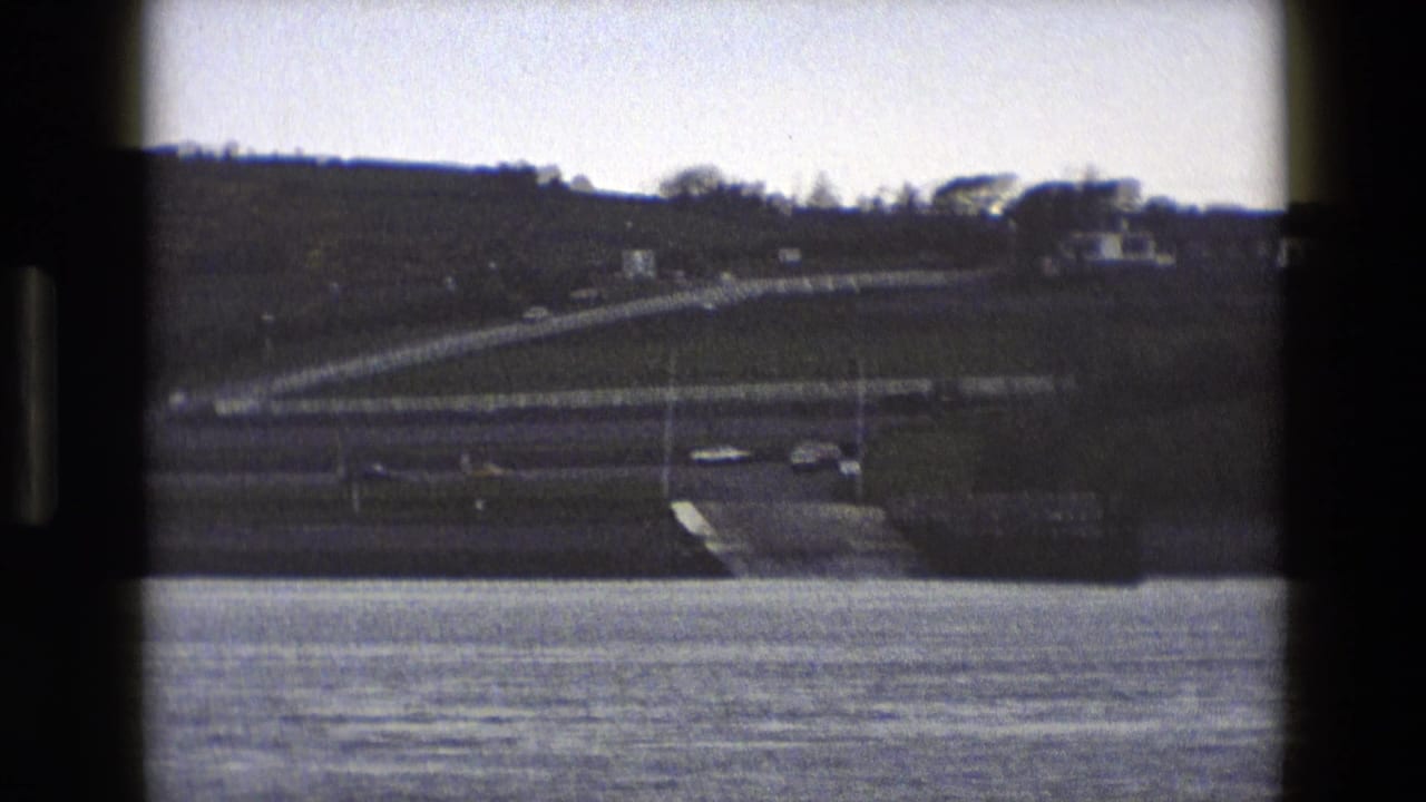 Cars move along a curving road above rippled water, with fencing and grassy hillside behind.