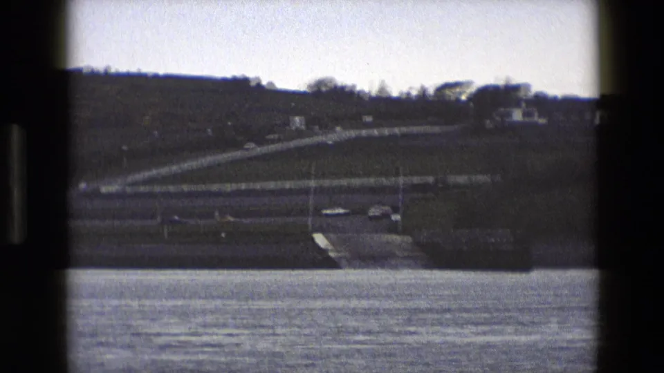 1969 England 8mm frame showing cars on a curving roadside ramp viewed across water, with white fencing and a grassy slope beyond.