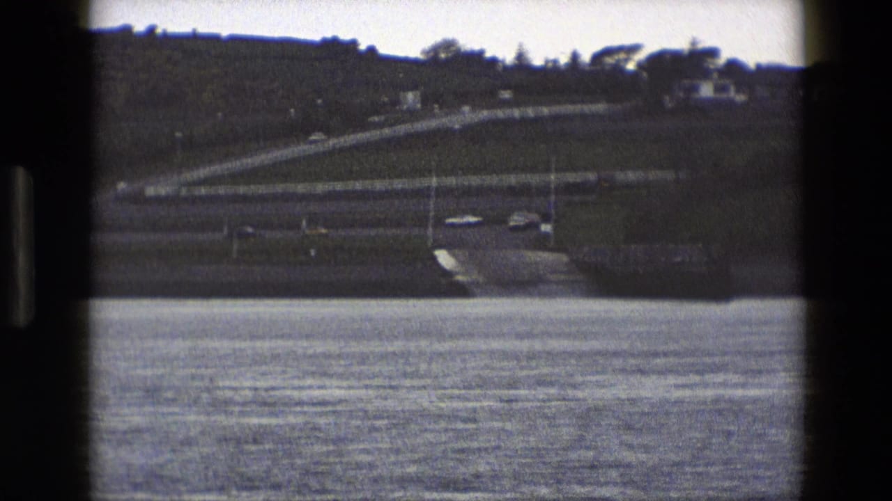 A road ramp and several cars are seen beyond water, with hillside fencing and distant houses.