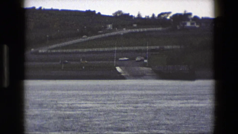 1969 England 8mm view of cars near an exit ramp across a body of water, framed by dark film edges and hillside fencing.