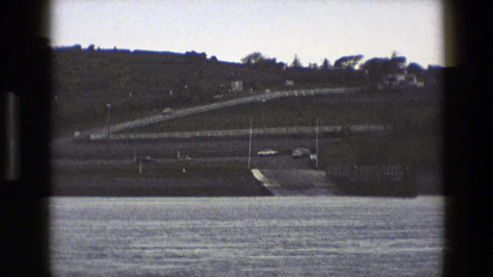 1969 England home-movie frame of roadside traffic near water, showing a curved ramp, cars, fencing, and a grassy hillside.