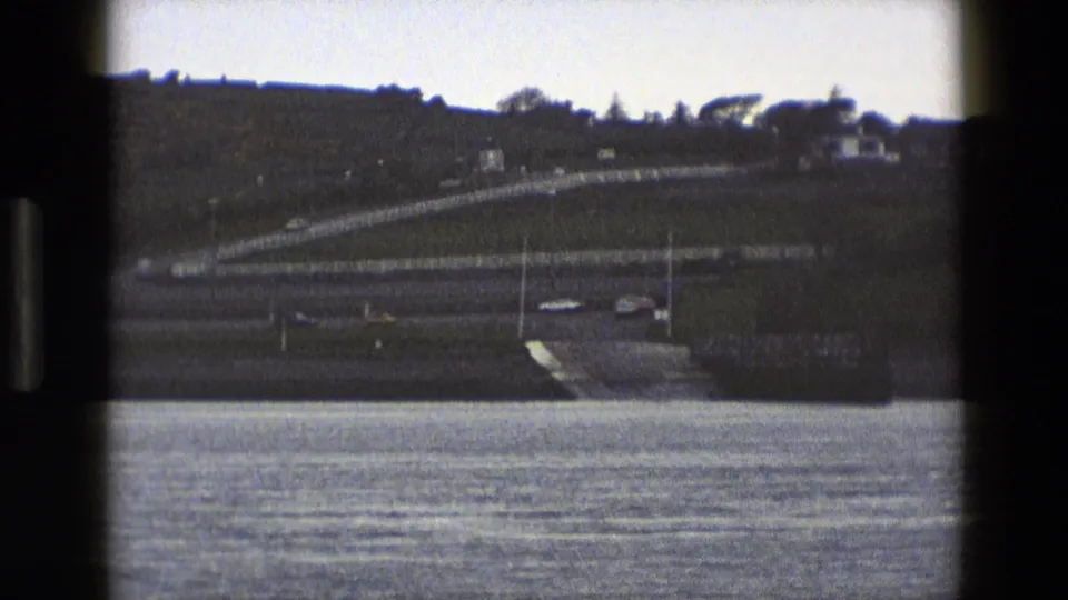 1969 England 8mm frame showing a waterfront view toward traffic on a ramp and road below a grassy hill with distant houses.