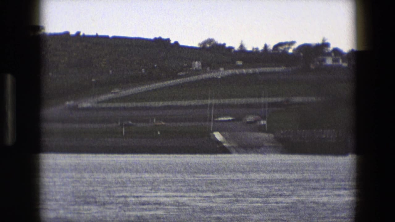 Calm water in front of a distant hillside with a road and small pale building.