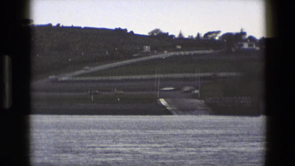 1969 England 8mm frame showing calm water, a dark hillside, a winding road, and a small pale building near the shore.
