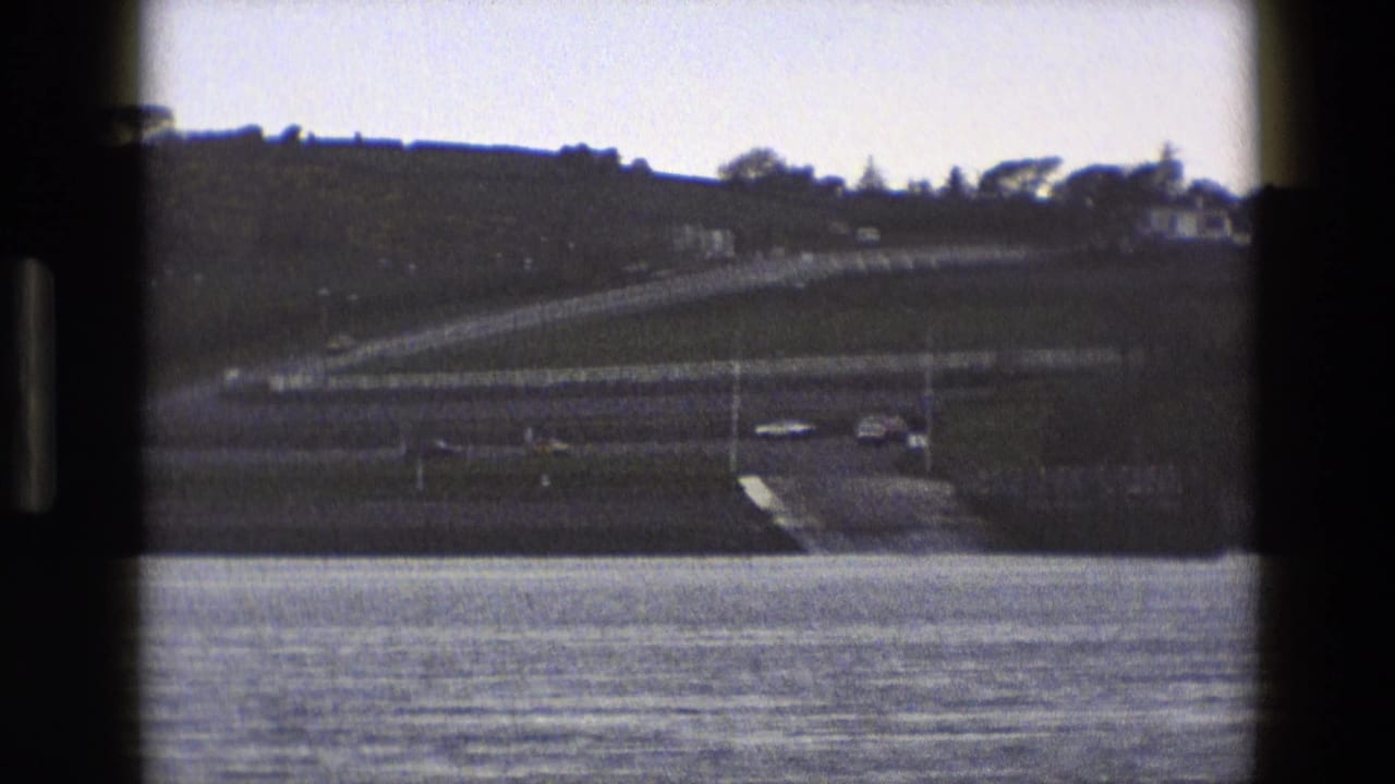1969 England 8mm frame of a calm water foreground, hillside road, and small pale structures on the far bank.