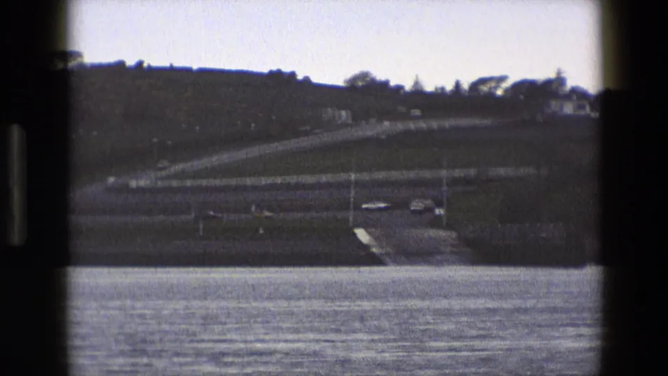 1969 England 8mm frame of a calm water foreground, hillside road, and small pale structures on the far bank.