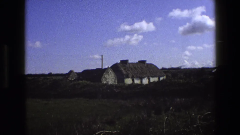 1969 England 8mm frame showing a whitewashed thatched cottage in open grass below a bright blue sky with scattered clouds.