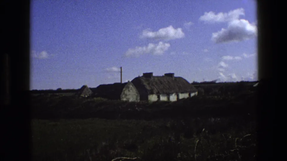 1969 England 8mm frame with a thatched rural cottage, nearby small structure, dark hedgerow, and blue sky with clouds.
