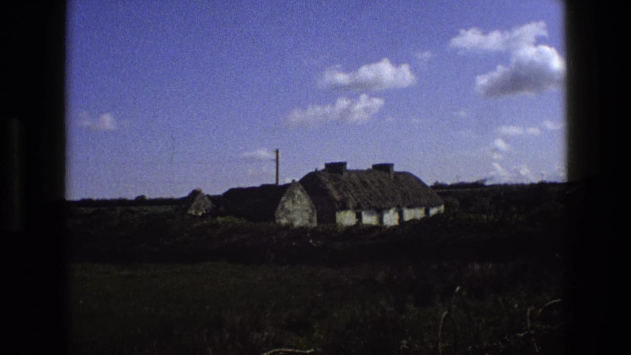 1969 England home-movie frame showing a distant thatched cottage, low hedges, open fields, and scattered clouds.