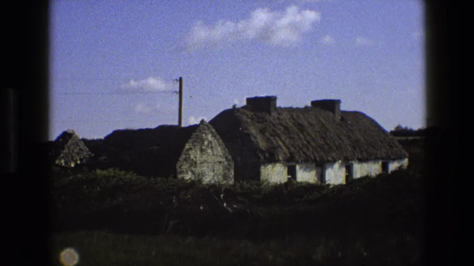 1969 England home-movie frame showing a thatched cottage, white walls, utility pole, overhead wire, and rural vegetation.