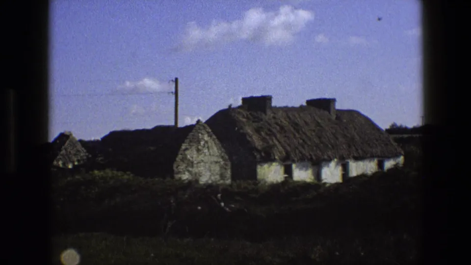 1969 England frame of a rural thatched cottage with a utility pole, overhead wire, hedge, and blue sky within dark film edges.