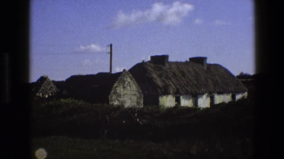 1969 England home-movie frame showing a whitewashed thatched cottage, hedgerow, utility pole, and scattered clouds.