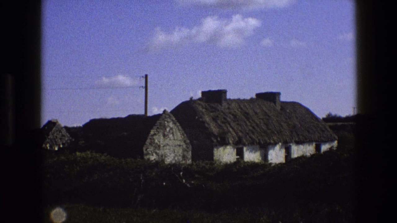 1969 England frame of a traditional thatched cottage viewed from outside, with hedges and a utility pole nearby.