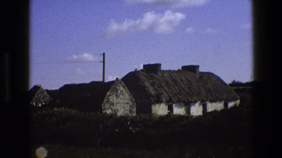 1969 England frame of a traditional thatched cottage viewed from outside, with hedges and a utility pole nearby.