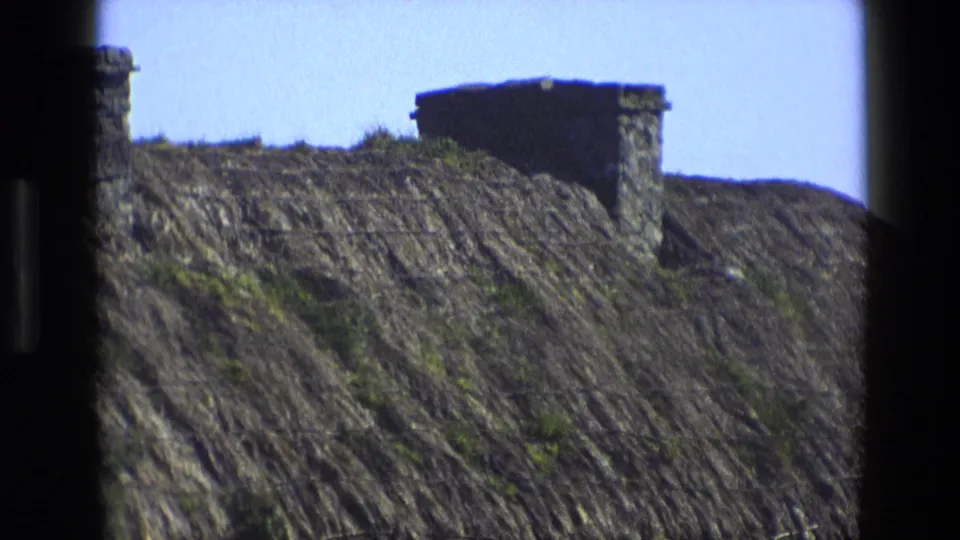 1969 England frame showing a sloped thatched roof, stone chimney, wire mesh, and bright sky within dark film borders.