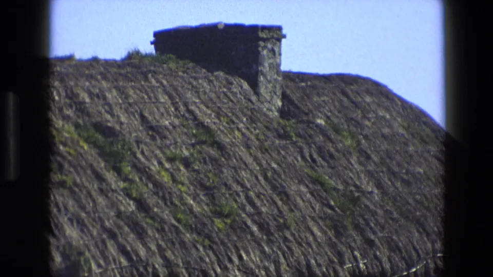 1969 England home-movie frame showing a close view of a thatched cottage roof and stone chimney under clear sky.