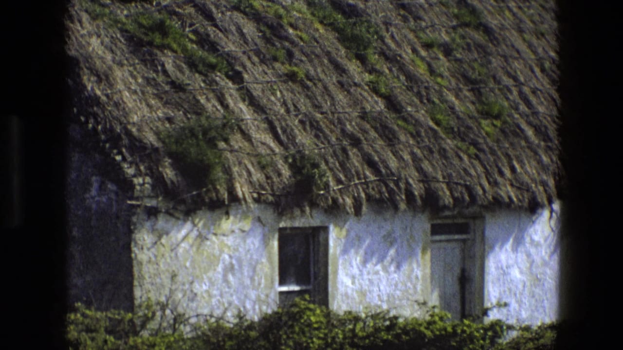 1969 England rural cottage exterior with whitewashed wall, heavy thatch, moss growth, small window, door, and dark vertical foreground edge.