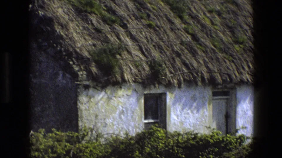 1969 England 8mm frame showing the cottage lower wall, window, door, dense hedging, and textured thatch under dark foreground edges.