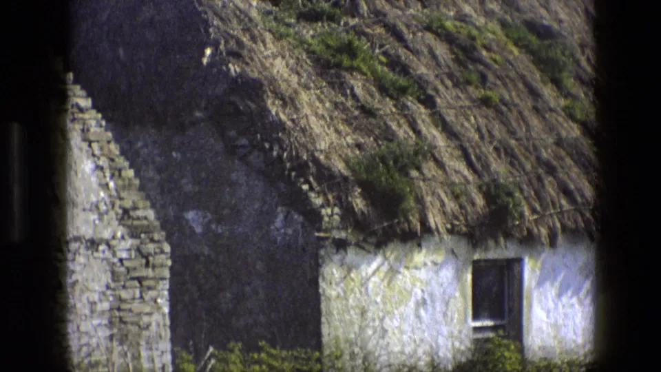 1969 England 8mm view of a cottage end with stone gable detail, whitewashed wall, mossy thatch, small window, and surrounding greenery.