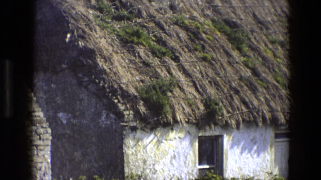 1969 8mm view of a whitewashed thatched cottage, partly obscured by a dark foreground edge, with a stone wall and grass below.