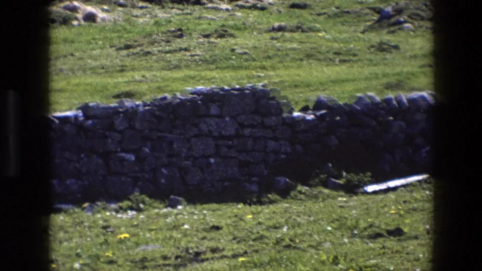 1969 England 8mm frame of a rural field, low stone wall, rocks, and grass partly obscured by dark window-frame edges.