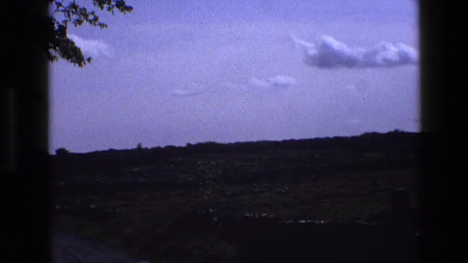 1969 England 8mm frame looking toward a pale sky, distant dark field edge, clouds, and tree leaves at the upper border.