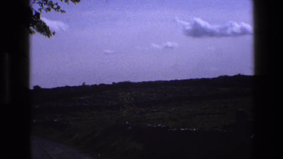 1969 England home-movie view of green countryside and clouds, framed by dark edges and tree branches at upper left.