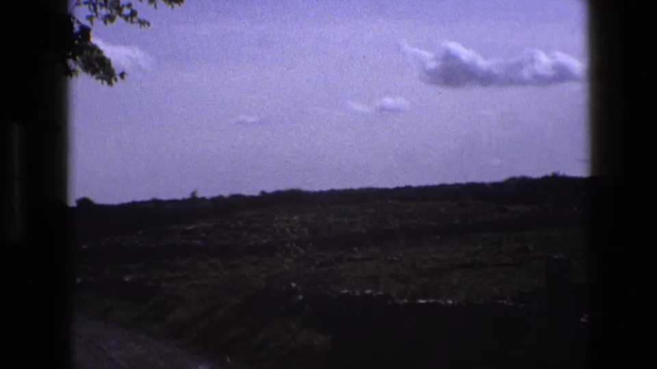 1969 England home-movie frame showing green fields, a low horizon, clouds, and dark window-like edges during travel.