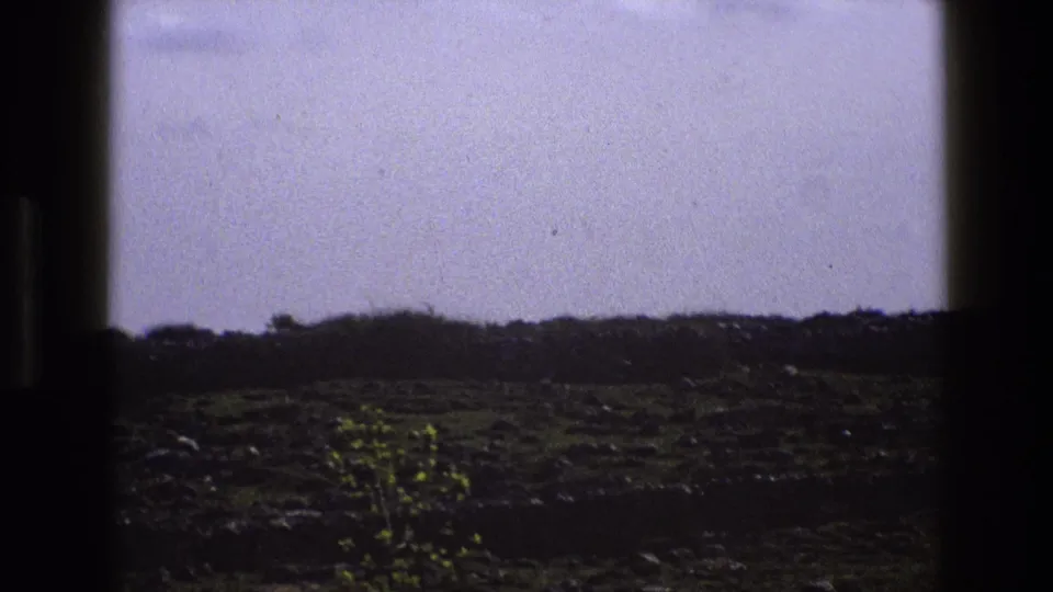 1969 England home-movie frame showing a rocky rural field and low horizon viewed through dark window-frame edges.