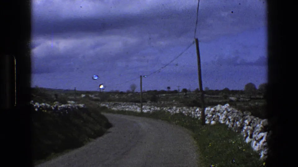 1969 England rural road curving through open fields with dry-stone walls, utility poles, and an overcast sky.