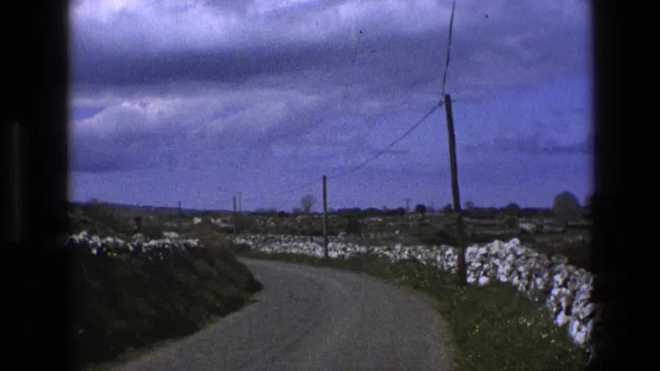 1969 England countryside frame with a narrow road, dry-stone wall, fields, poles, and visible film gate edges.