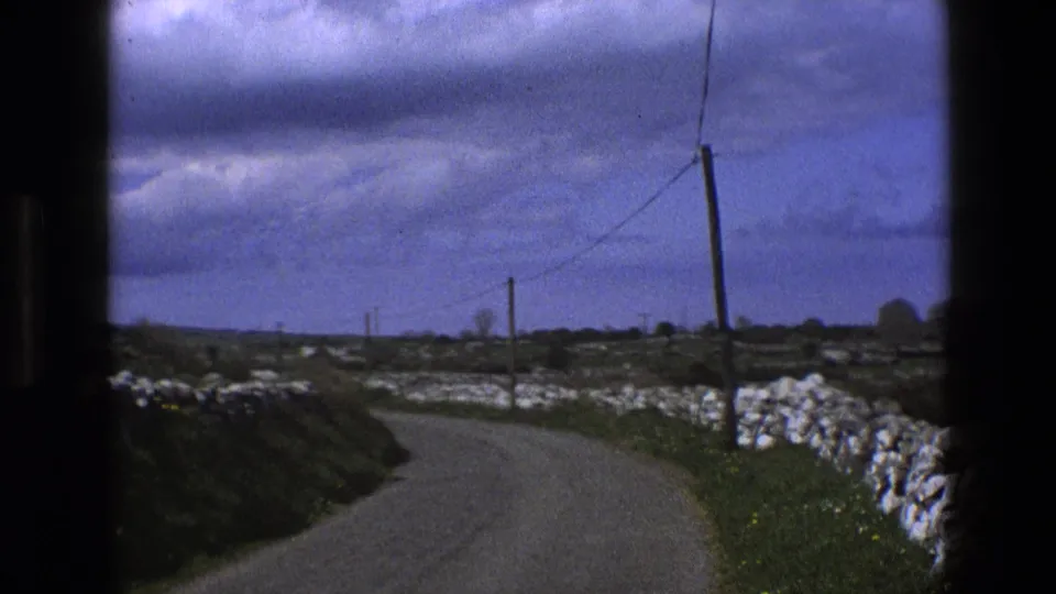 1969 England 8mm view from a moving vehicle on a curving country road with white stone walls, fields, and telephone wires.