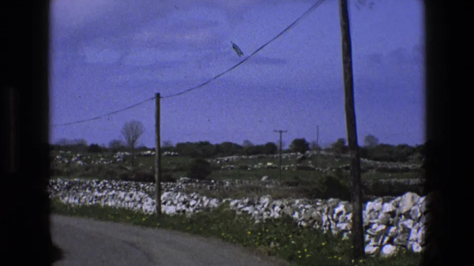 1969 England 8mm frame showing roadside stone walls, green fields, utility poles, and a pale blue cloudy sky.