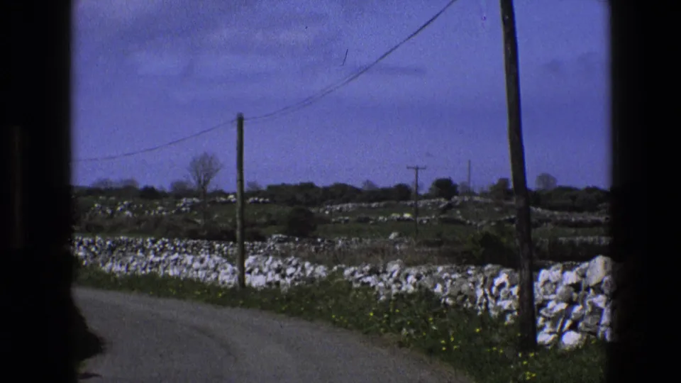 1969 England rural road scene with low white stone walls, green fields, bare trees, and telephone poles along the lane.