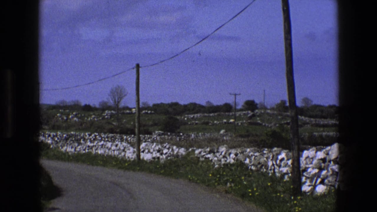 1969 England home-movie frame of a rural road curving past dry-stone walls, fields, wooden poles, and overhead lines seen from a vehicle.