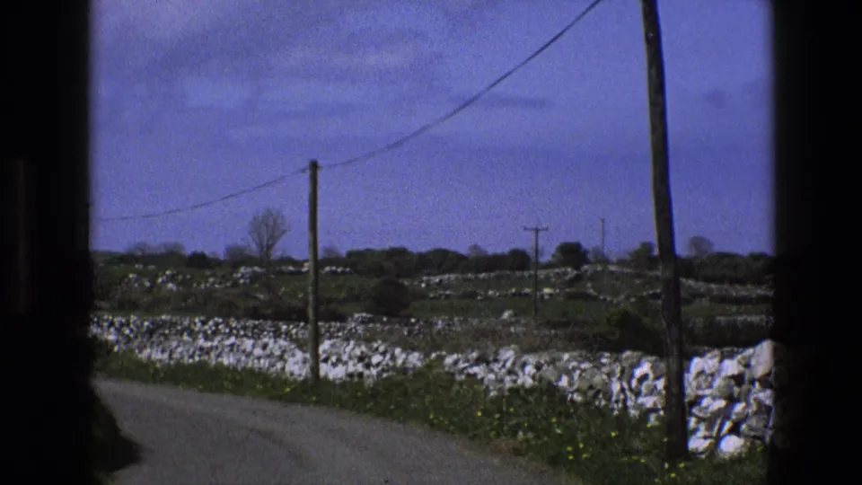 Blue-cast 1969 England 8mm frame of a narrow rural lane, dry-stone walls, fields, utility poles, and overhead lines from a car window.