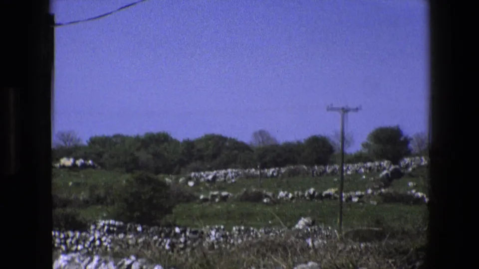 1969 England countryside view with green fields, dry-stone walls, hedgerows, a wooden utility pole, and overhead wires in soft 8mm color.
