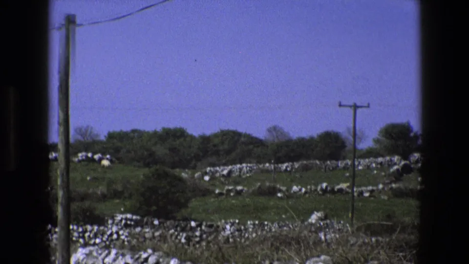 1969 England hillside landscape with layered stone walls, green pasture, a telephone pole, overhead wire, trees, and blue sky.
