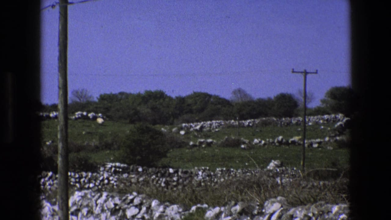 1969 England 8mm frame of pastureland seen through dark framing, with dry-stone walls, shrubs, a utility pole, and open blue sky.