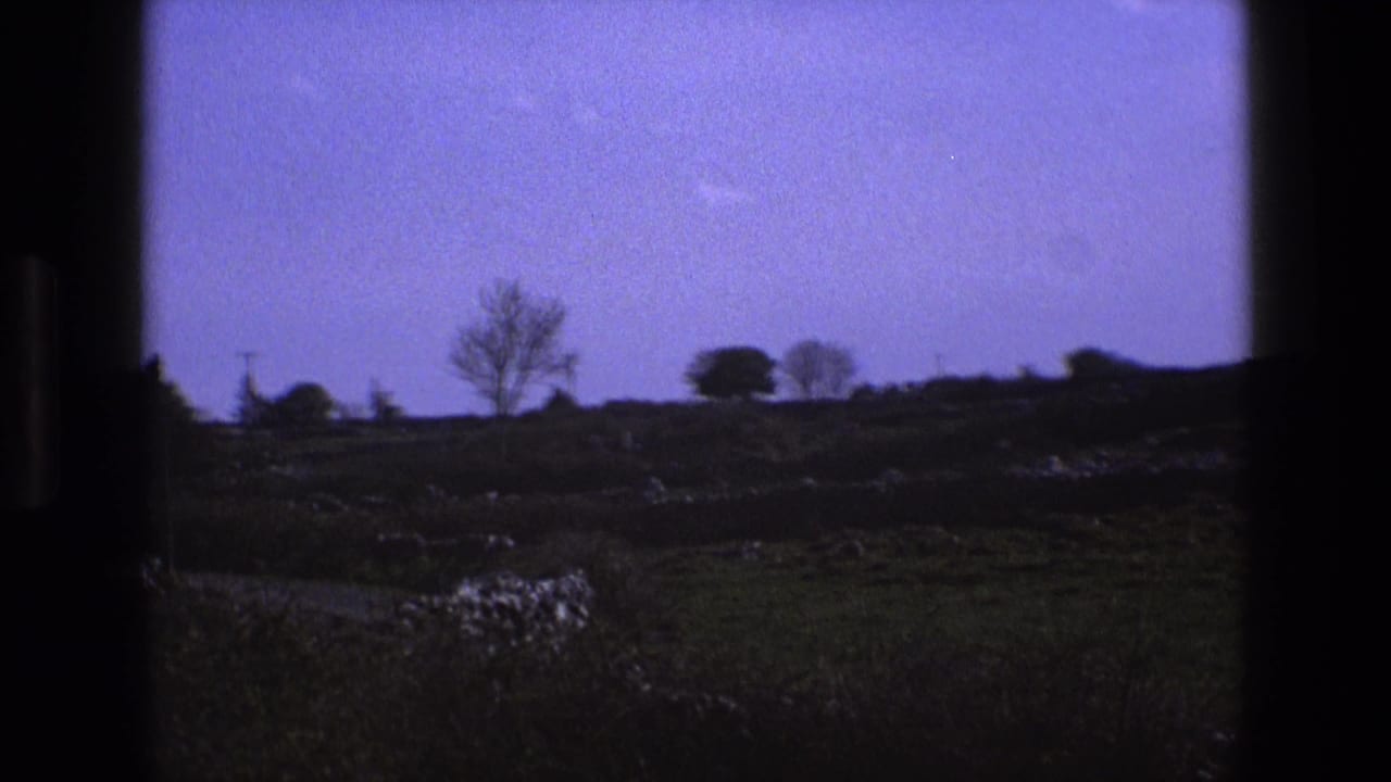 1969 England home-movie frame showing open countryside through a window, with fields, low stone walls, distant trees, and a purple-blue sky.