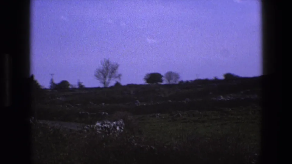 1969 England home-movie frame showing open countryside through a window, with fields, low stone walls, distant trees, and a purple-blue sky.